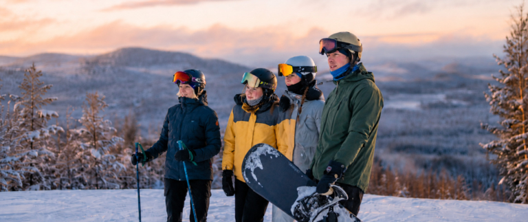 Fyra personer i skid- och snowboardutrustning står på en snötäckt höjd vid Hassela och blickar ut över ett vinterlandskap i kvällsljus.